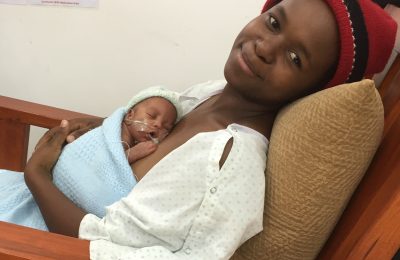 Picture of a women sitting in a chair with her little baby sleeping on her abdomen, making 'skin-to-skin'. The mother is smiling peacefully